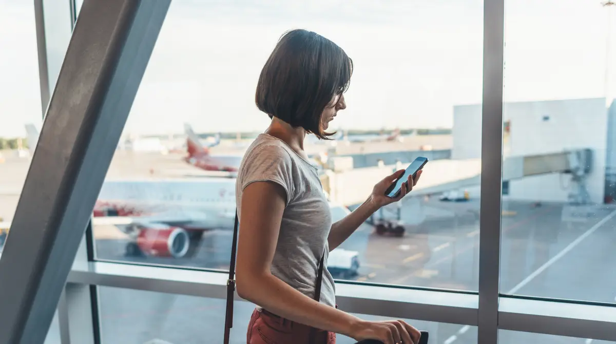Udhëtar duke mbajtur telefonin në një terminal aeroporti para dritares së nisjeve