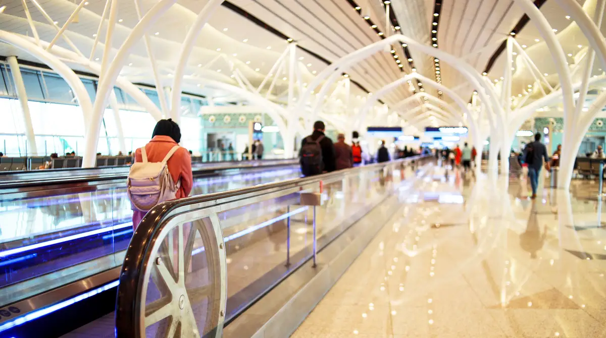 Udhëtarë në një shirit lëvizës brenda një terminali modern aeroporti me kolona të bardha të degëzuara